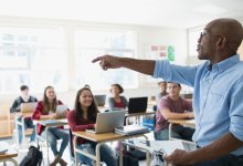 Photo of a male teacher calling on a student in a classroom filled with smiling, seated high schoolers